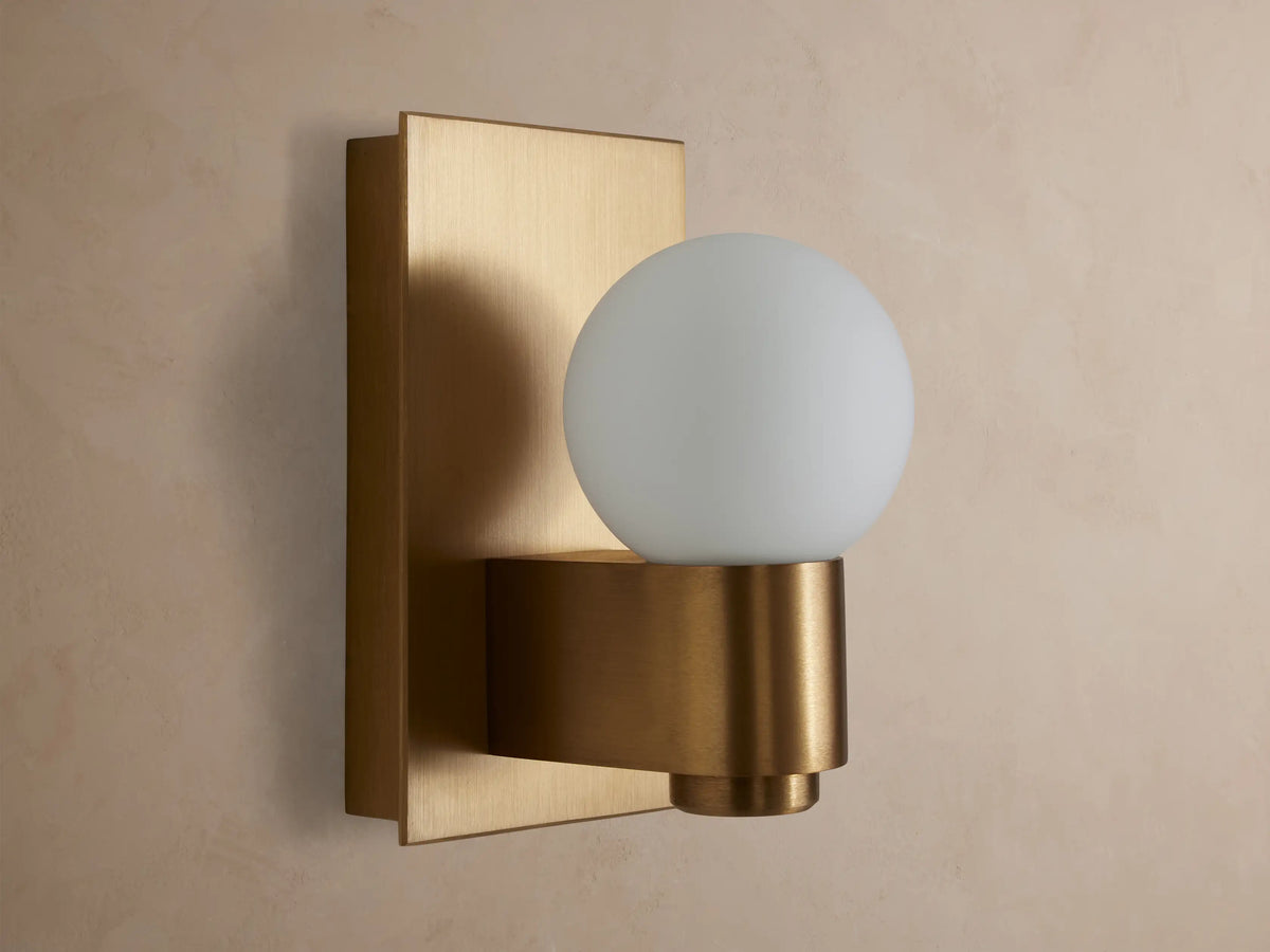 Illuminated Casen Sconce with a glowing mouth-blown solid opal glass globe on a satin brass fixture.