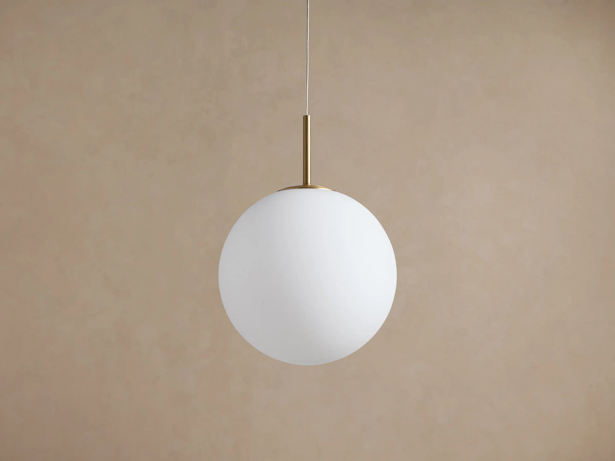 Casen Pendant: Illuminated white opal glass globe suspended from a satin brass rod and thin wire against a warm wall.