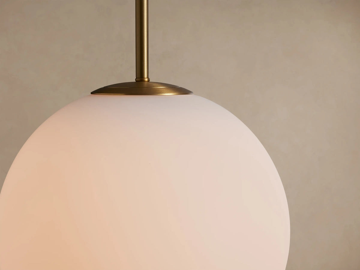 Casen Pendant: Spherical white opal glass globe light hanging from a satin brass rod and thin silver wire against a plain wall.