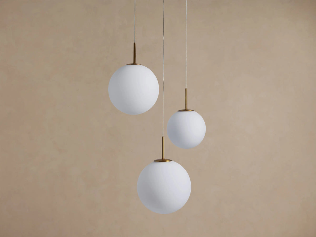Casen Round Chandelier: Three white opal glass globes with satin brass stems, suspended at varying heights against a beige wall.
