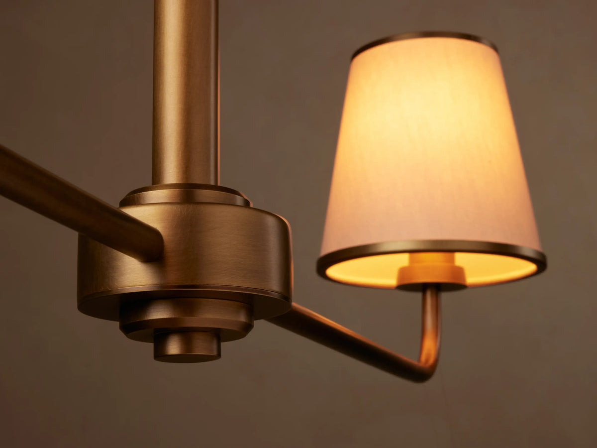 Adria 2-Light Chandelier: horizontal bronze bar, two conical cream fabric shades with gold trim, hanging against a plain beige wall.