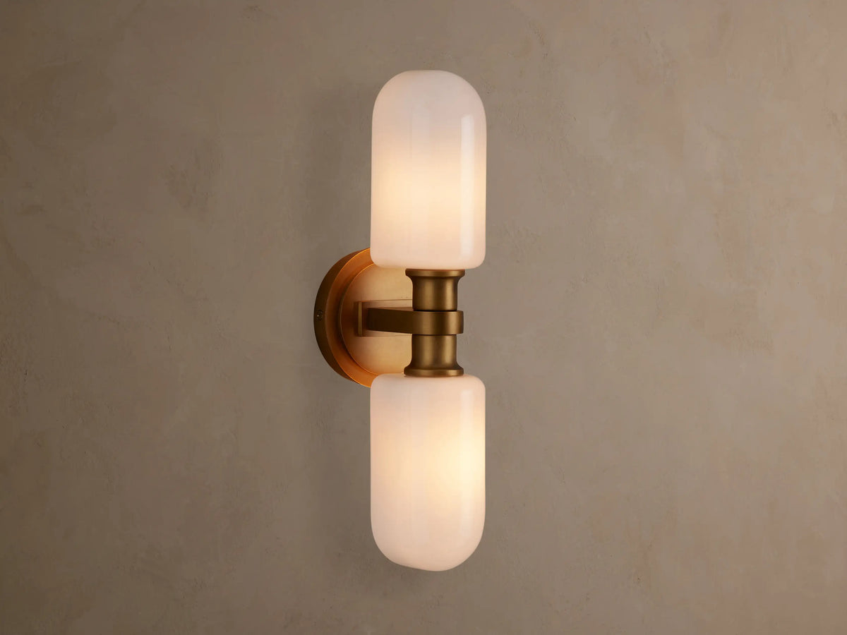 Zayd Double Sconce, vertically mounted, with two illuminated white cylindrical glass shades and brass-toned hardware on a textured wall.