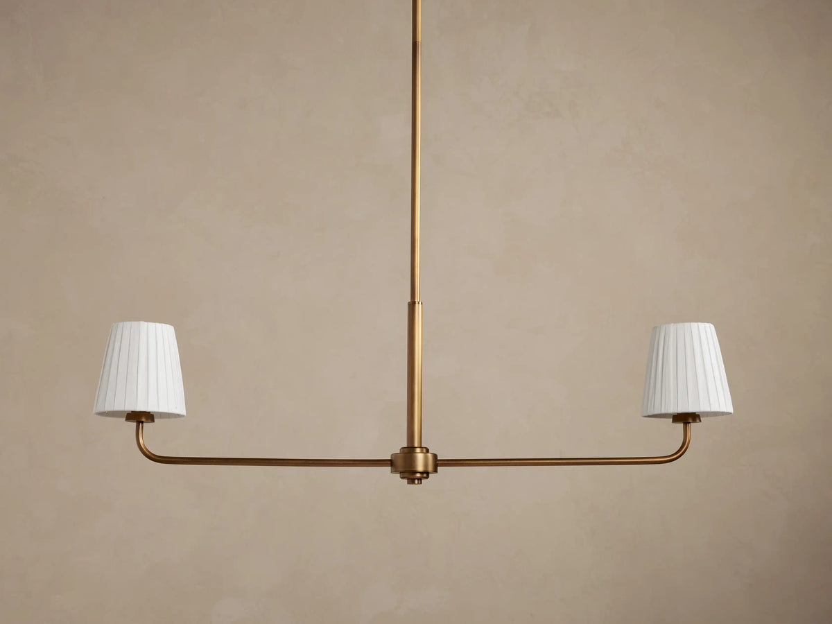 Adria 2-Light Chandelier with brass-toned frame, central rod, and two pleated off-white shades illuminated against a brown wall.