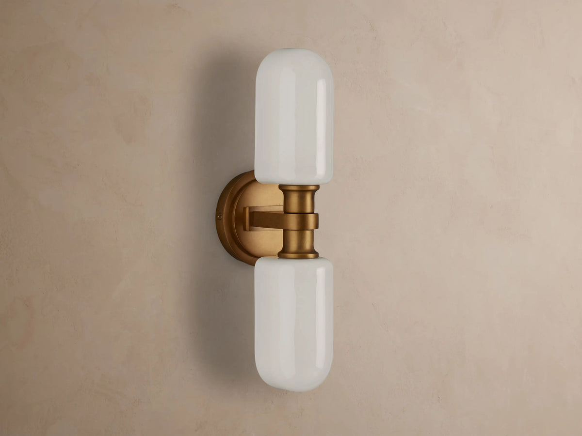 Zayd Double Sconce, vertically mounted, with two illuminated white cylindrical glass shades and brass-toned hardware on a textured wall.