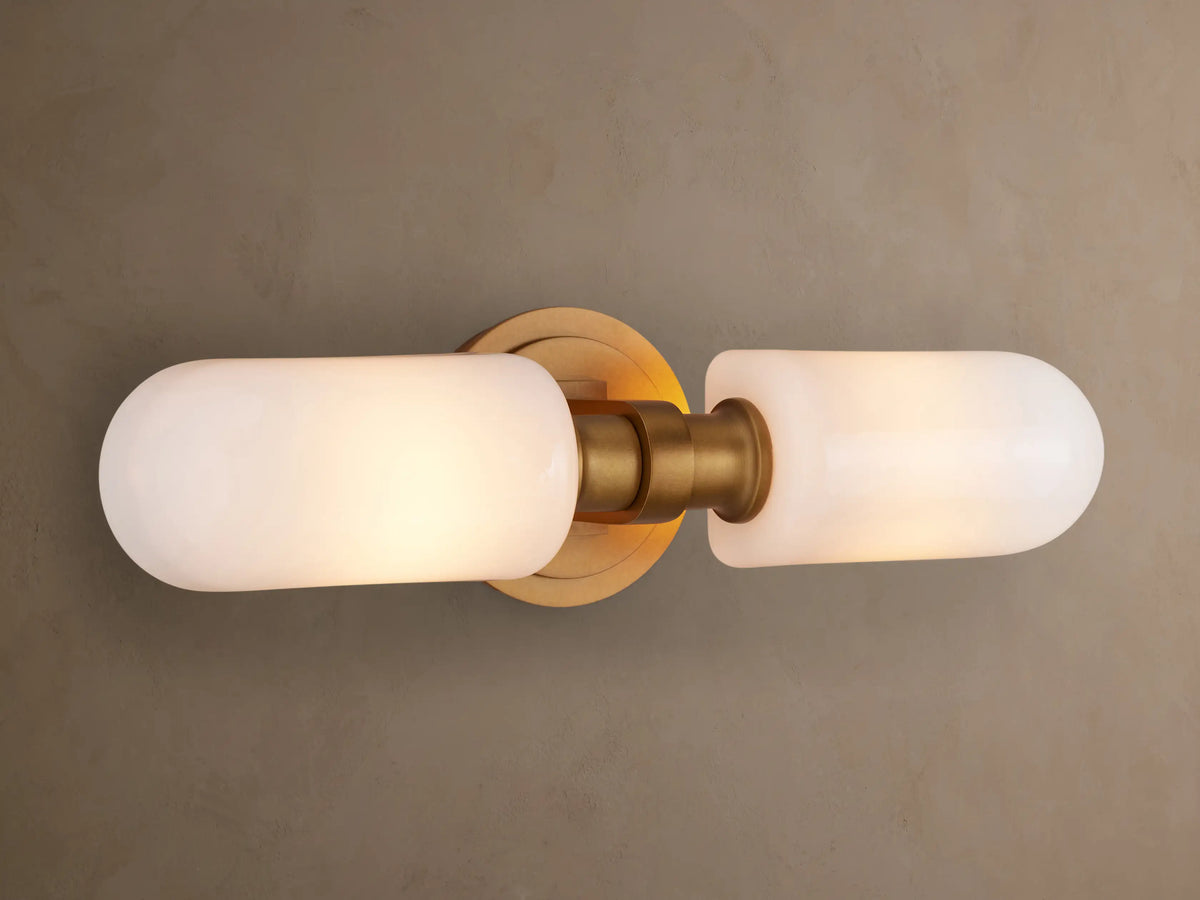 Zayd Double Sconce: Vertical brushed brass fixture with two illuminated frosted white glass shades, on a gray wall.