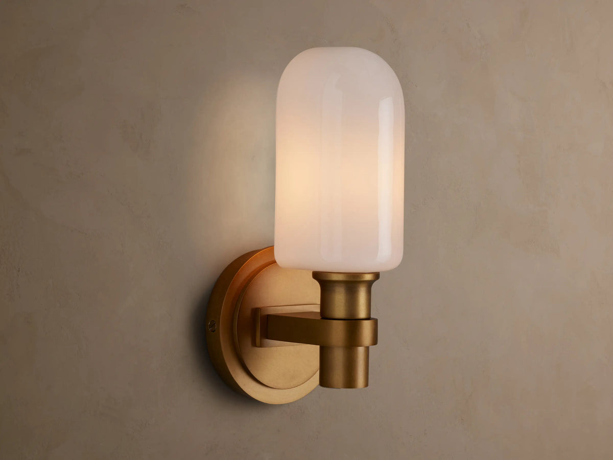 Zayd Sconce, an upright brass wall light with a glowing, rounded opal glass shade mounted on a textured beige wall.