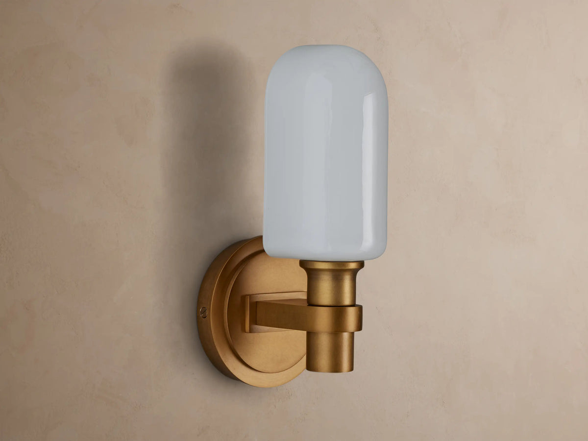 Zayd Sconce, an upright brass wall light with a glowing, rounded opal glass shade mounted on a textured beige wall.