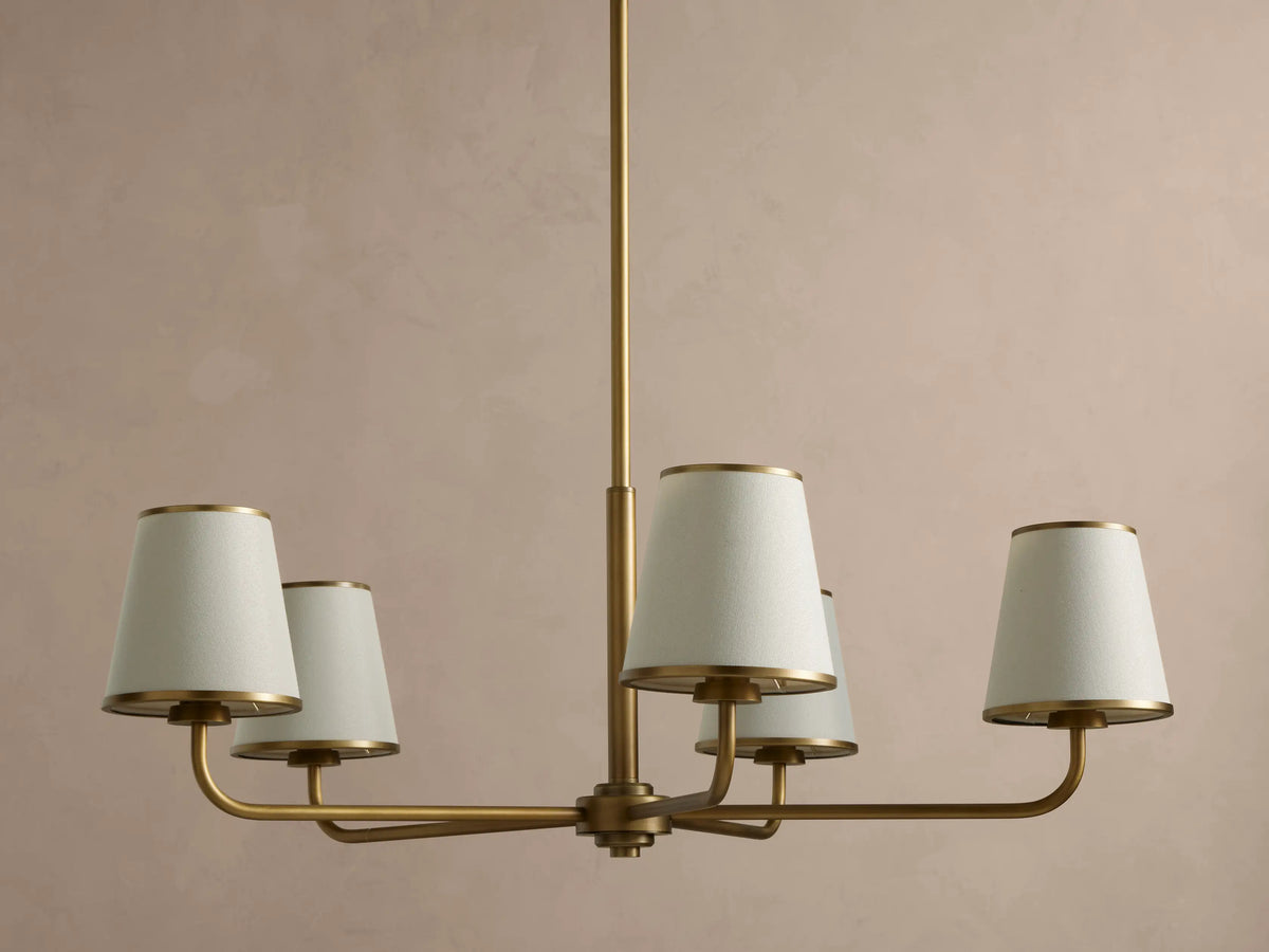 Adria Round Chandelier: brass fixture with 5 cream conical shades, gold metal trim, hanging from a rod on a beige wall.