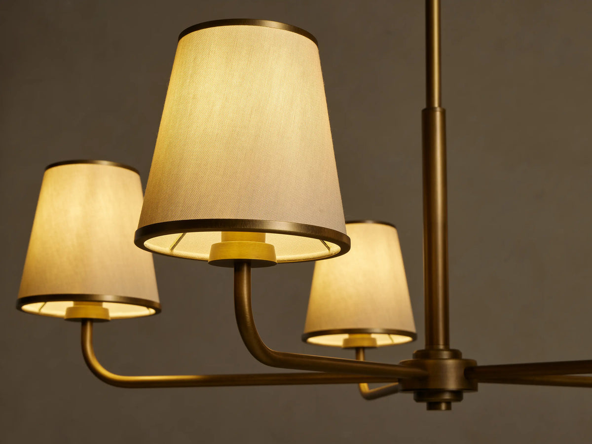 Adria Round Chandelier, close-up. Three lit cream conical fabric shades with dark metal trim on a brushed brass frame.