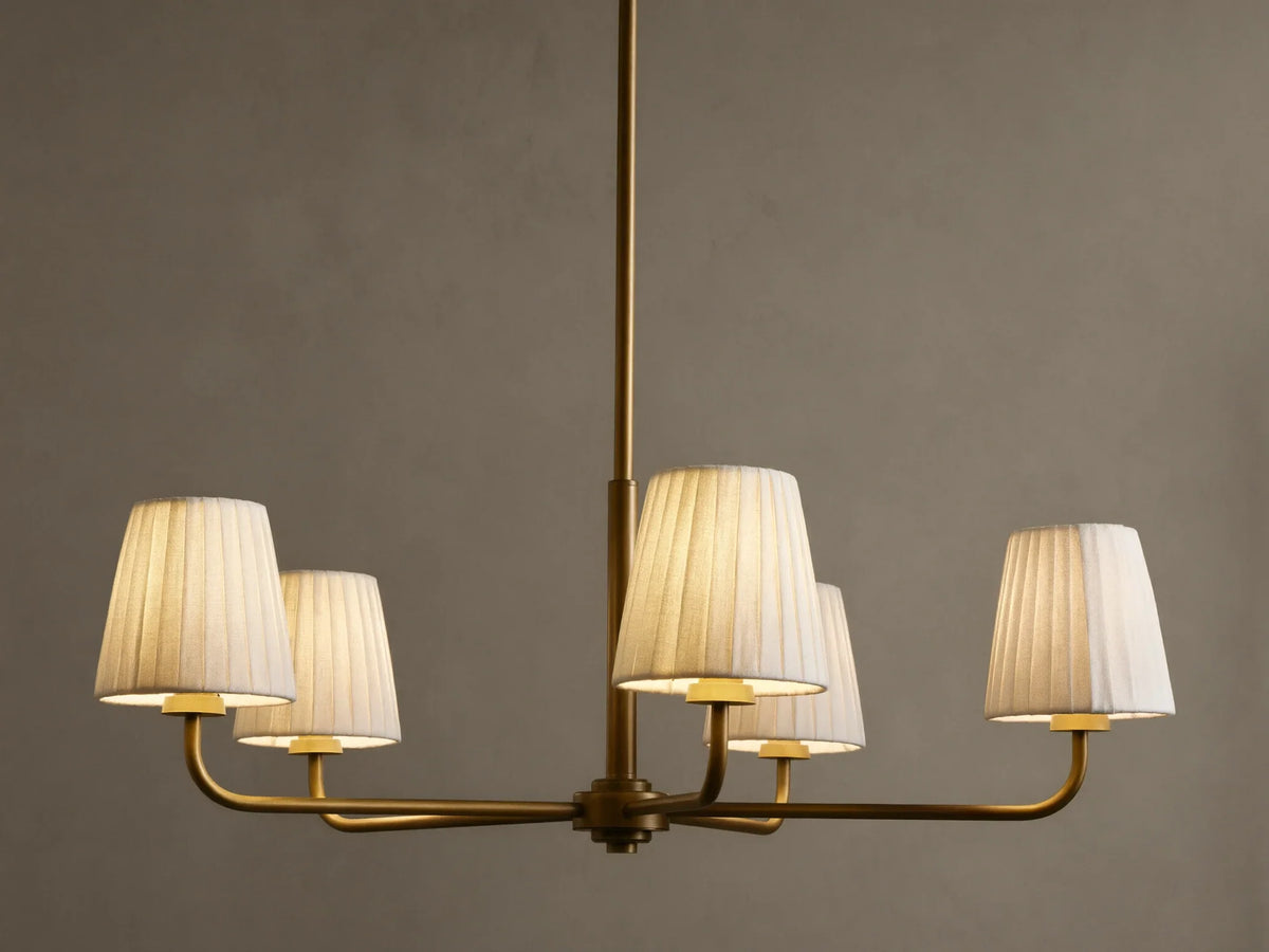 Adria Round Chandelier with five illuminated pleated white shades on a brass-finished frame, hanging against a dark wall.