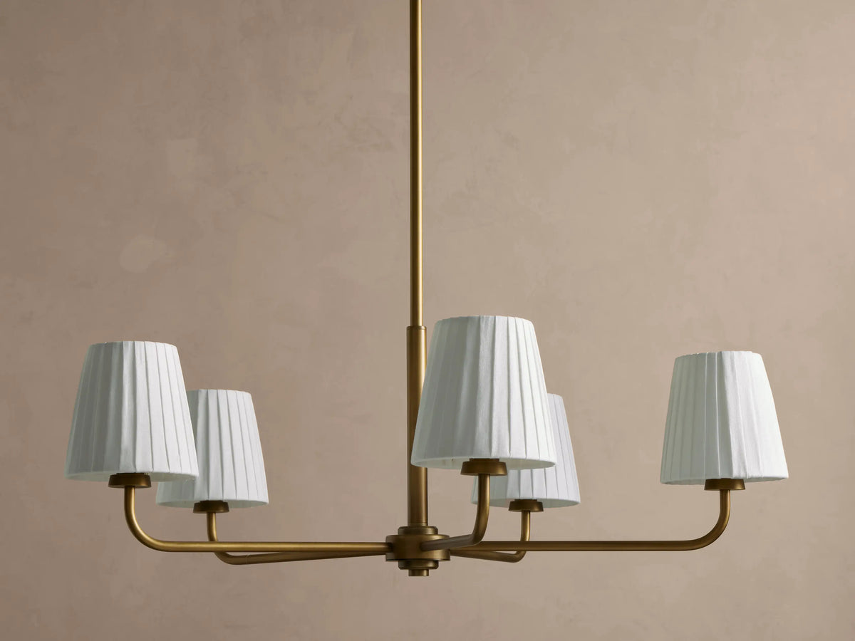 Adria Round Chandelier: golden iron frame with five white pleated shades, hanging against a light beige wall.
