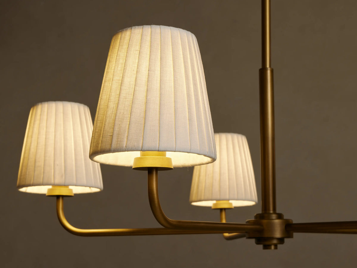Adria Round Chandelier; three glowing pleated off-white shades on an antique brass metal frame, seen against a dark background.