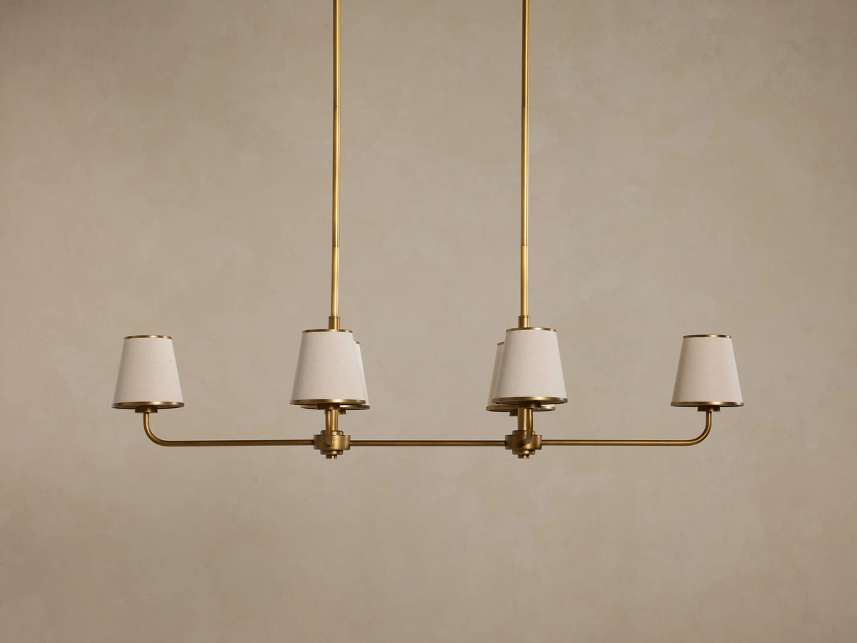 Adria Linear Chandelier with four glowing, tapered beige fabric shades trimmed in brass, suspended by two brass rods.