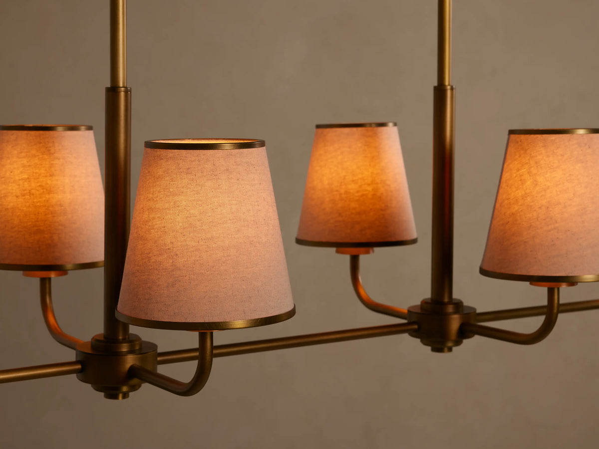 Adria Linear Chandelier: four tapered cream shades on a horizontal brass bar, suspended by two vertical rods against a light wall.