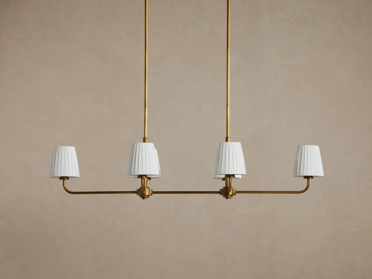 Adria Linear Chandelier with 4 illuminated pleated white shades on a horizontal brass bar, hanging from 2 brass rods.