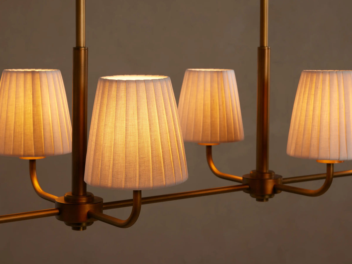 Adria Linear Chandelier, gold finish, with four white pleated shades, hanging from two suspension rods against a neutral wall.