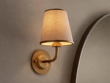 Adria Sconce: A wall-mounted, brass-toned sconce with a lit, conical, light beige fabric shade, casting a warm glow on a textured wall.