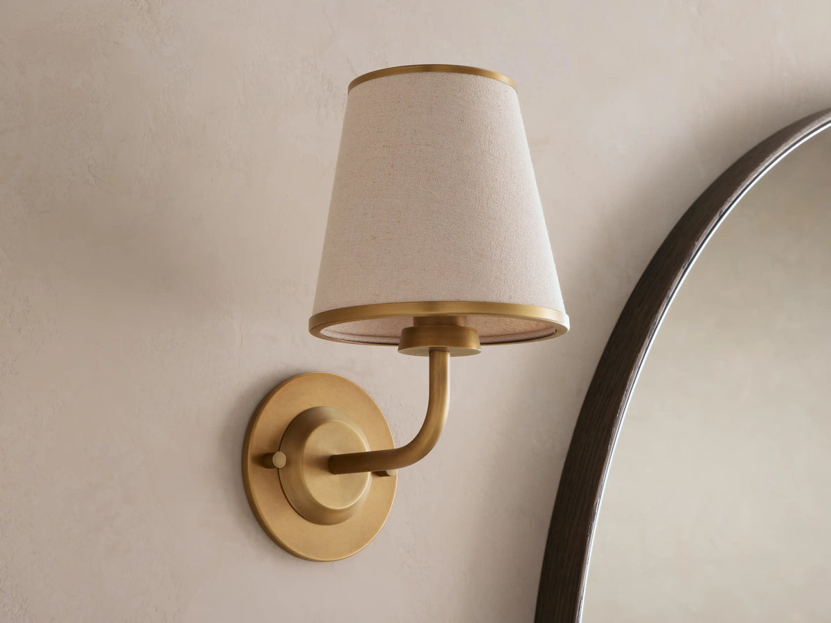 Adria Sconce: A wall-mounted, brass-toned sconce with a lit, conical, light beige fabric shade, casting a warm glow on a textured wall.