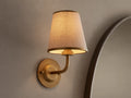 Adria Sconce: A wall-mounted, brass-toned sconce with a lit, conical, light beige fabric shade, casting a warm glow on a textured wall.