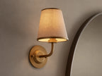 Adria Sconce: A wall-mounted, brass-toned sconce with a lit, conical, light beige fabric shade, casting a warm glow on a textured wall.