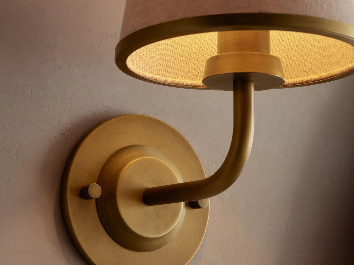 Adria Sconce with brass base and arm, and a tapered cream fabric shade with brass trim, mounted on a light wall next to a mirror.