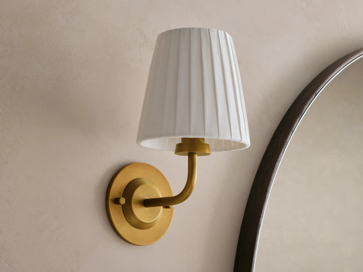 Adria Sconce: brass fixture, pleated beige shade, warm light on a textured wall beside a dark wood mirror.