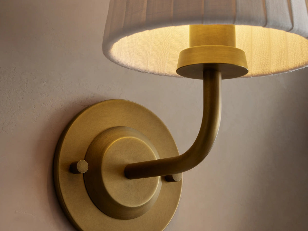 Adria Sconce featuring a round brass base, curved brass arm, and a white pleated fabric shade, mounted on a light wall.