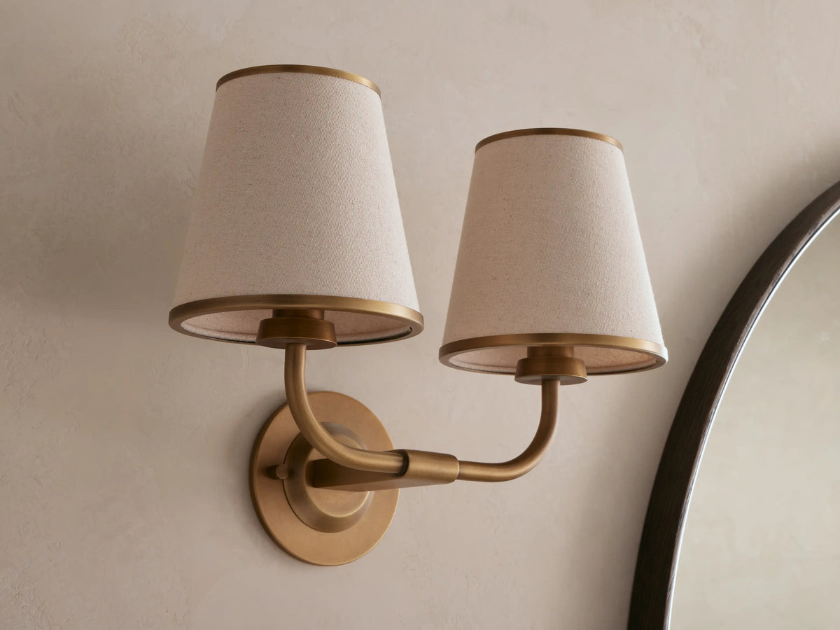 Adria Double Sconce with two illuminated light beige fabric shades, golden metal frame, mounted on a light wall next to a mirror.