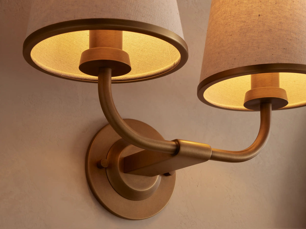 Adria Double Sconce with two cream fabric shades, brass-toned iron frame, mounted on a light wall next to a dark mirror.