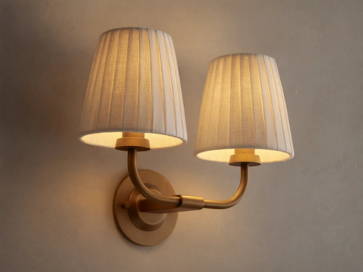 Adria Double Sconce with two lit off-white pleated shades on a brass-toned iron base, mounted on a wall next to a mirror.