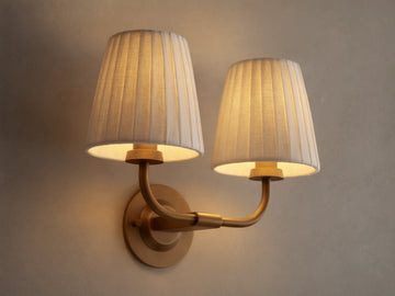 Adria Double Sconce with two lit off-white pleated shades on a brass-toned iron base, mounted on a wall next to a mirror.