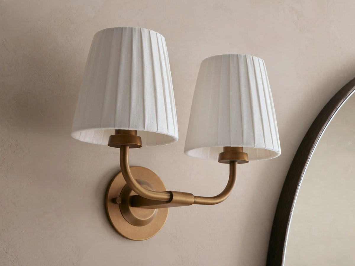 Adria Double Sconce with two lit off-white pleated shades on a brass-toned iron base, mounted on a wall next to a mirror.
