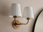 Adria Double Sconce with two lit off-white pleated shades on a brass-toned iron base, mounted on a wall next to a mirror.