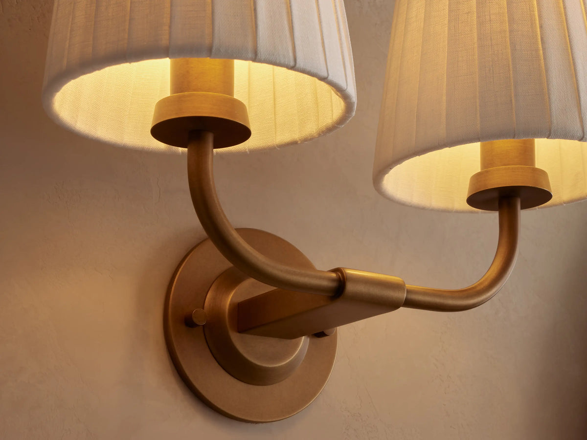 Adria Double Sconce: Two white pleated shades on a curved brass-finished iron frame, mounted on a beige wall. Partial mirror right.