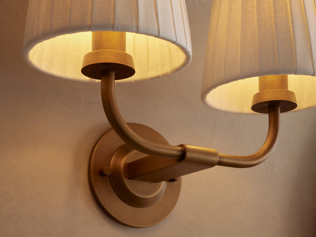 Adria Double Sconce: Two white pleated shades on a curved brass-finished iron frame, mounted on a beige wall. Partial mirror right.