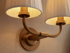 Adria Double Sconce: Two white pleated shades on a curved brass-finished iron frame, mounted on a beige wall. Partial mirror right.