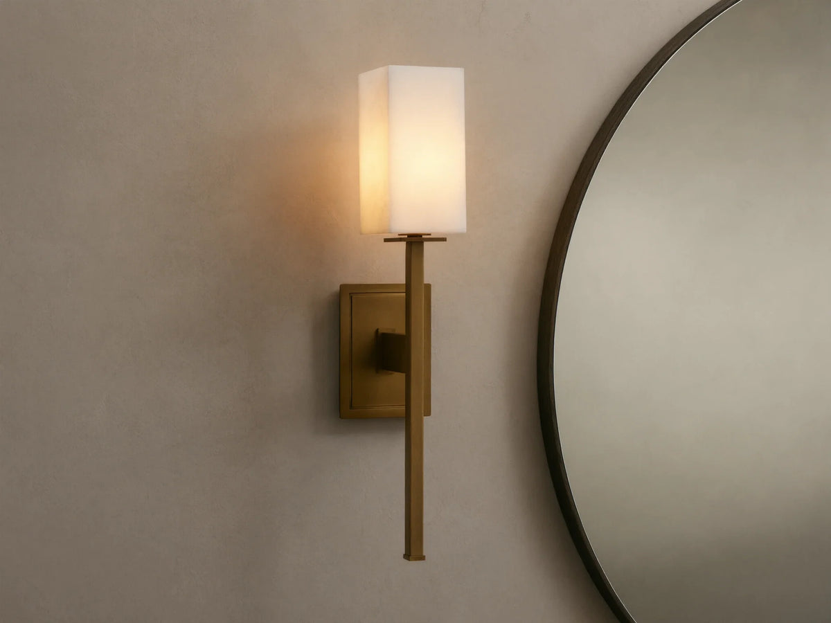 Cinda Sconce, a vertical brass finish wall light with a glowing rectangular stone shade, mounted on a tan wall next to a mirror.