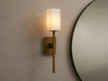 Cinda Sconce, a vertical brass finish wall light with a glowing rectangular stone shade, mounted on a tan wall next to a mirror.