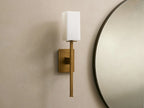Cinda Sconce, a vertical brass finish wall light with a glowing rectangular stone shade, mounted on a tan wall next to a mirror.