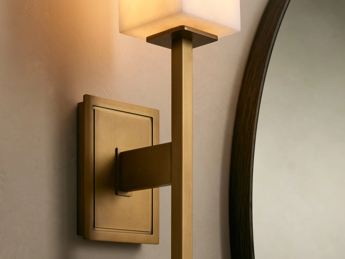 Cinda Sconce: Vertical brass-finished wall sconce with a rectangular white stone shade, mounted on a beige wall next to a round mirror.