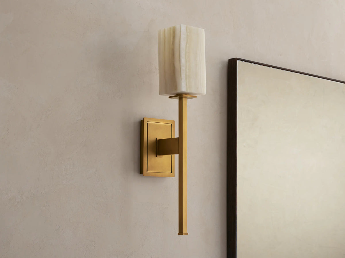 Cinda Sconce: Wall-mounted brass sconce with an illuminated rectangular natural stone shade next to a mirror on a beige wall.