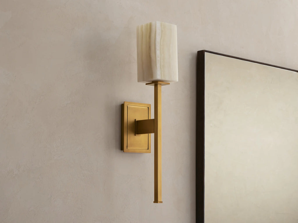 Cinda Sconce: Wall-mounted brass sconce with an illuminated rectangular natural stone shade next to a mirror on a beige wall.