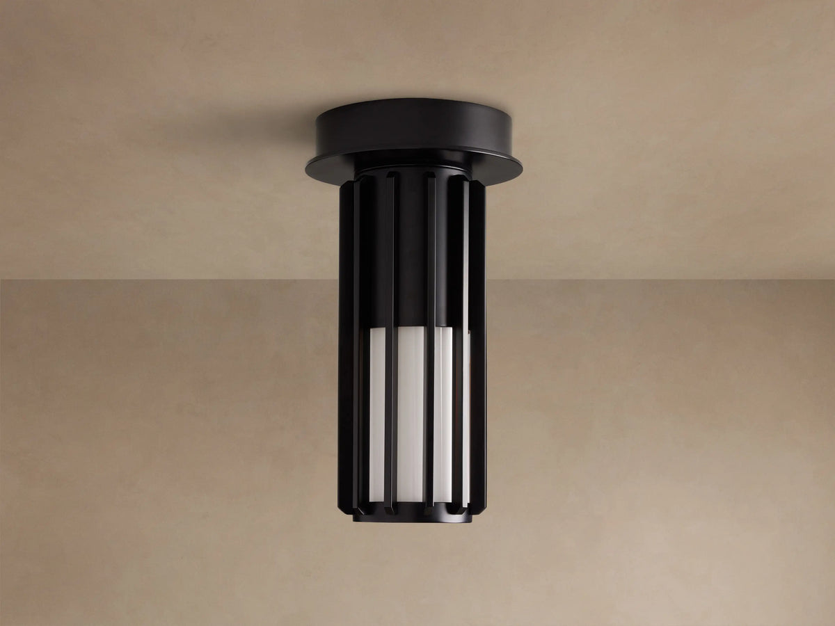 Matte black Gabbia Outdoor Spotlight Flush Mount, cylindrical with vertical slats, flush-mounted to a light brown ceiling. Inner milk glass panels glow.