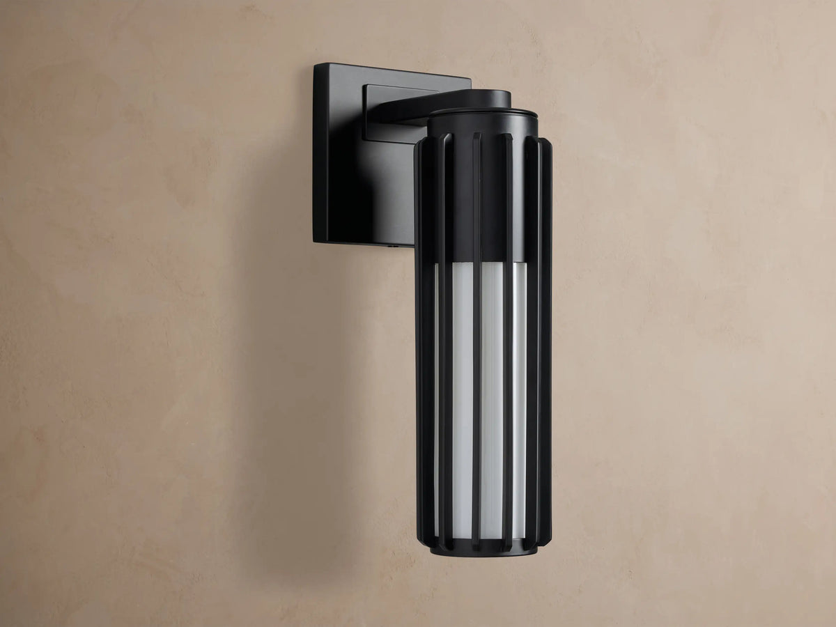 Matte black Gabbia Outdoor Sconce, cylindrical with vertical slats, illuminated warm milk glass, mounted on a beige wall.
