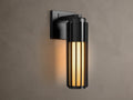 Matte black Gabbia Outdoor Sconce, cylindrical with vertical slats, illuminated warm milk glass, mounted on a beige wall.