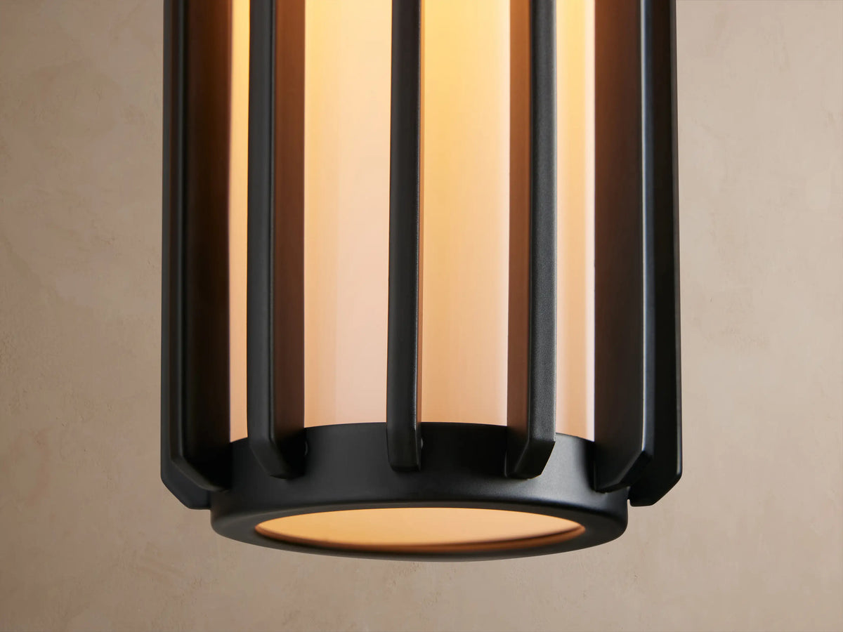 Matte black Gabbia Outdoor Sconce with a rectangular backplate, cylindrical ribbed body, and white milk glass, on a beige wall.