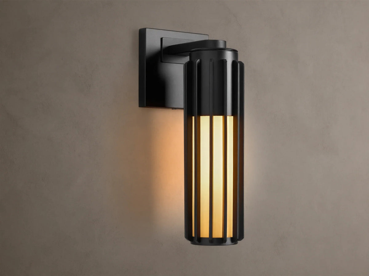 Matte black Gabbia Outdoor Sconce, cylindrical with vertical slats, illuminated warm milk glass, mounted on a beige wall.