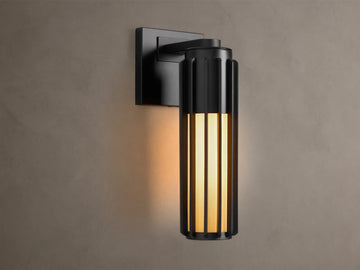 Matte black Gabbia Outdoor Sconce, cylindrical with vertical slats, illuminated warm milk glass, mounted on a beige wall.