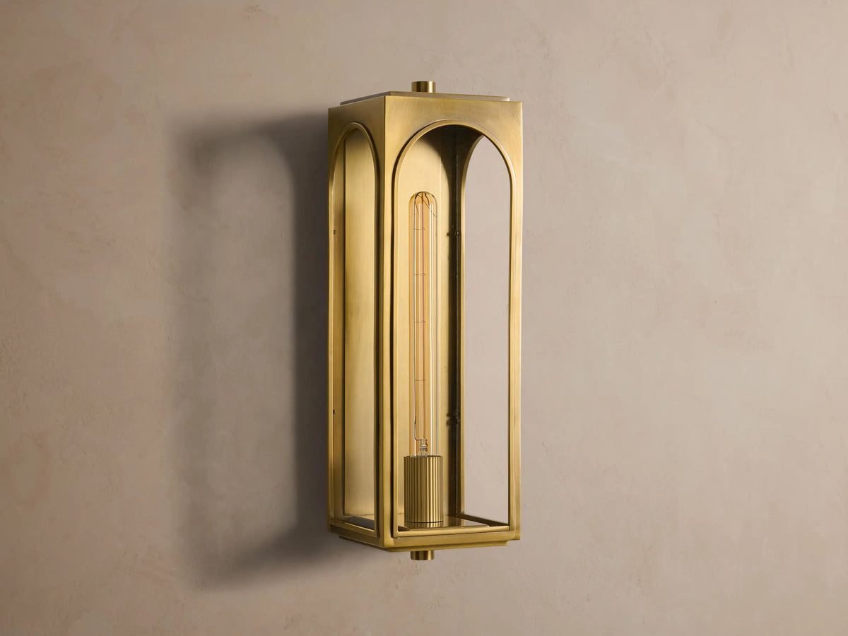 Brass Palladian Outdoor Sconce with arched glass panels, illuminating a warm Edison bulb. Mounted on a textured wall.
