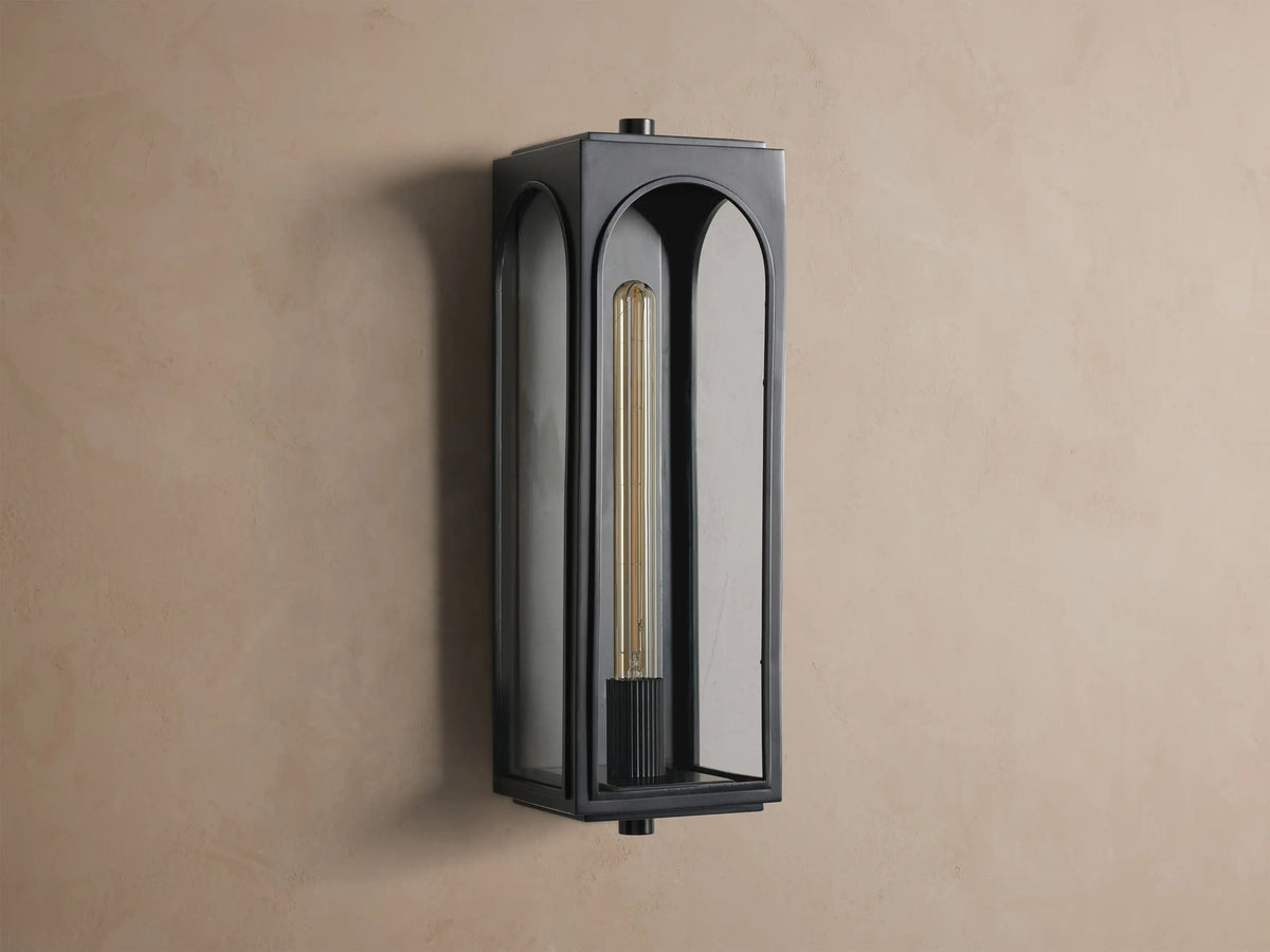 Black Palladian Outdoor Sconce, vertical, with arched glass panels and a warm glowing filament bulb, mounted on a tan wall.